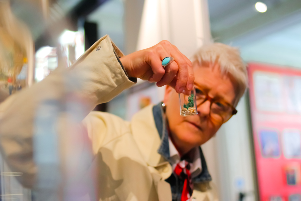 A person with short white hair and glasses examines a small object, possibly jewelry or an artifact, in their hand. They wear a light coat and turquoise ring, with displays and red walls in the blurred background.